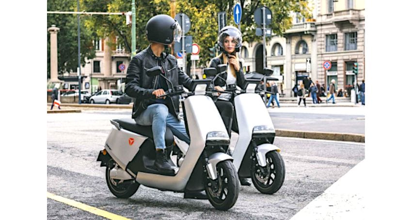 Two people wearing helmets riding electric scooters on a city street.