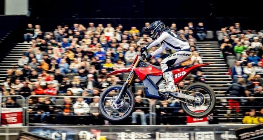 A motorcyclist in white and black gear is performing a jump mid-air on a red bike with a large crowd watching in the background.