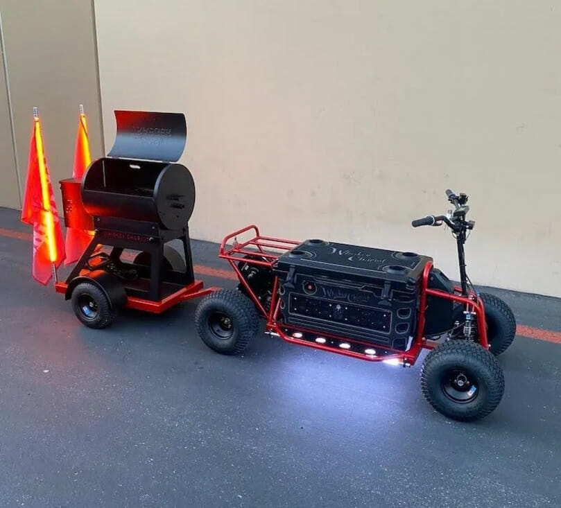 A red electric scooter towing a small black and red grill with two orange safety flags attached at the back. The scooter and grill are on a black asphalt surface in front of a beige wall.