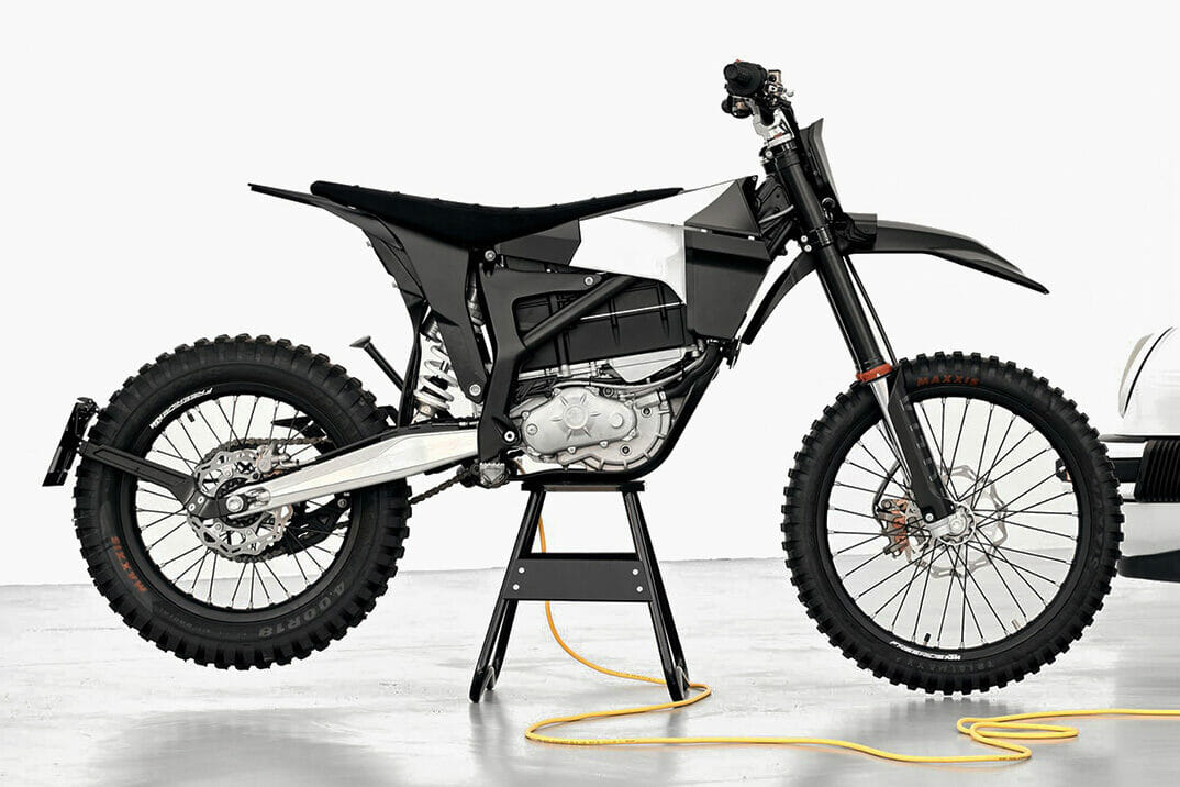 A black dirt bike is positioned on a stand indoors, with a yellow cable connected to it. The bike features knobby tires and a visible engine.