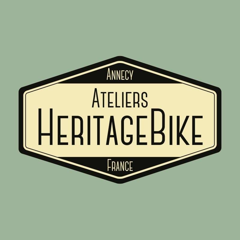 Logo of Ateliers HeritageBike featuring a beige hexagonal emblem with a black border. The text "Annecy Ateliers HeritageBike France" is prominently displayed in black. The background is muted green.