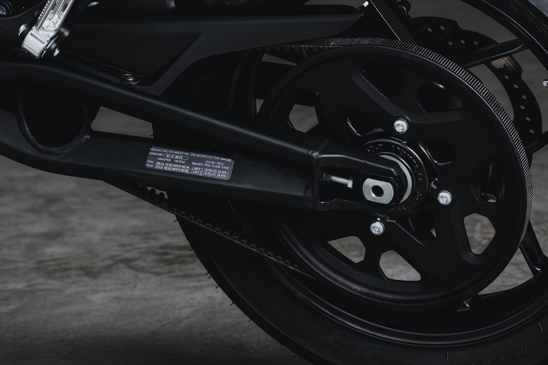 Close-up of the rear wheel and drive mechanism of a black motorcycle. The image shows the chain, sprocket, and part of the swingarm. The surface below the motorcycle is concrete.