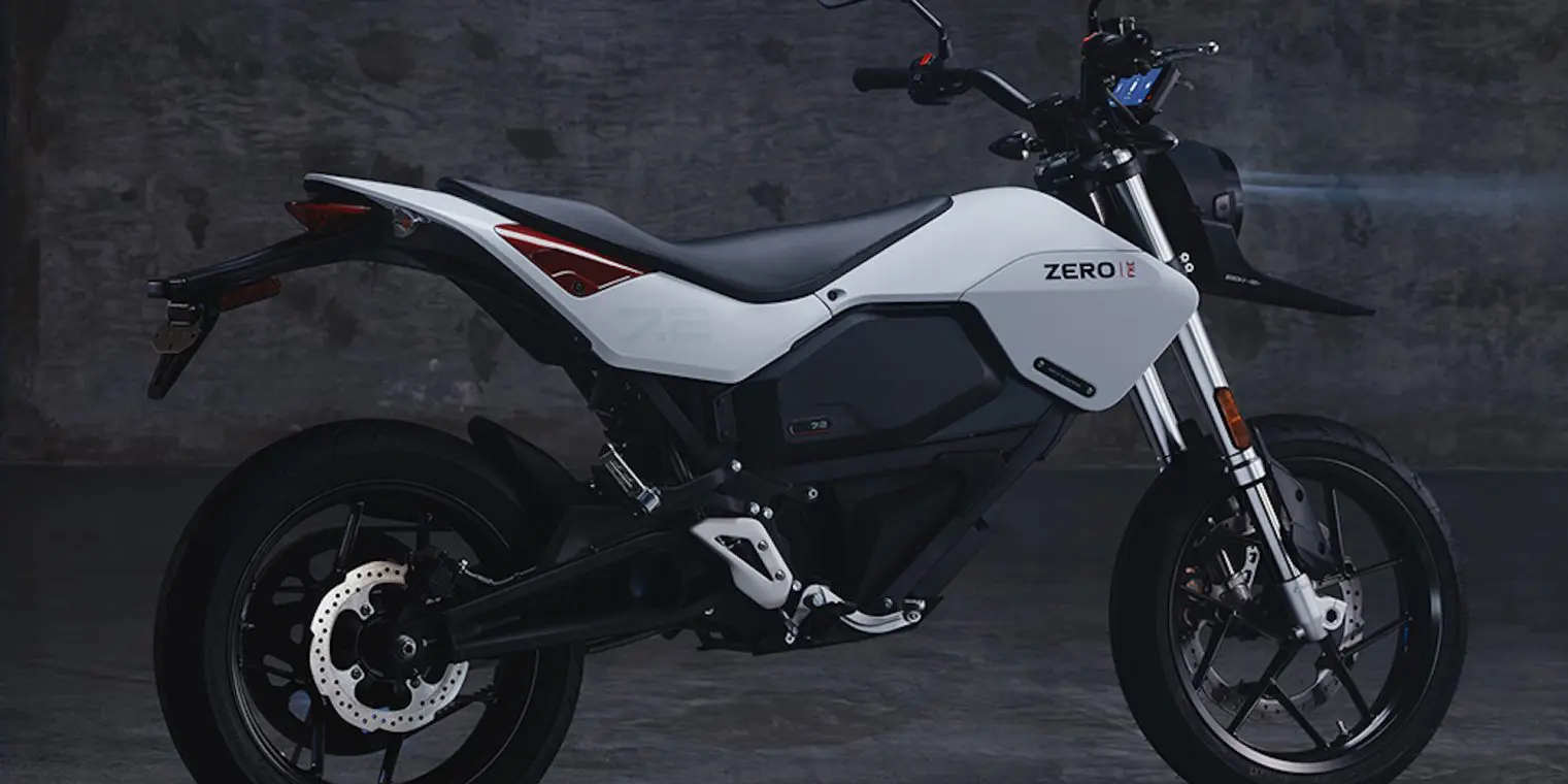 A white Zero electric motorcycle is parked on a dark, concrete floor. It features a minimalist design with black detailing and red accents near the rear.