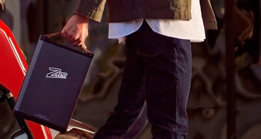 A person is carrying a black briefcase with a Zapp EV logo on it while walking, wearing denim jeans and a jacket with a white shirt underneath.