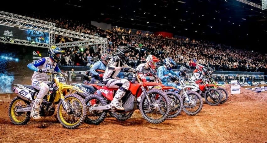 Motocross riders lined up at the start line in an indoor arena, with a crowd of spectators in the background.