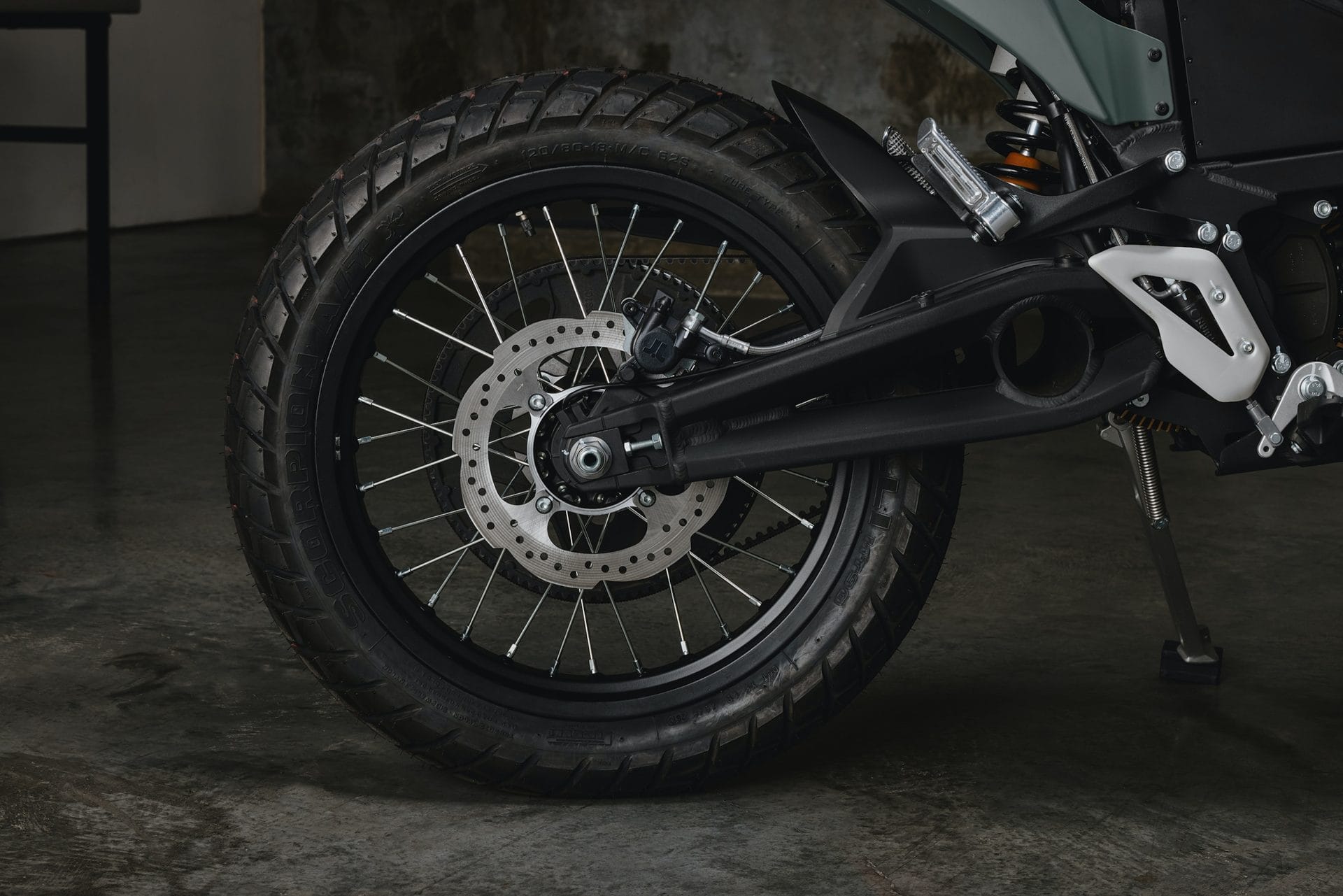 Close-up of a motorcycle's rear wheel and brake disc assembly, featuring a black tire, spokes, and a detailed view of the chain and suspension system on a textured concrete floor.