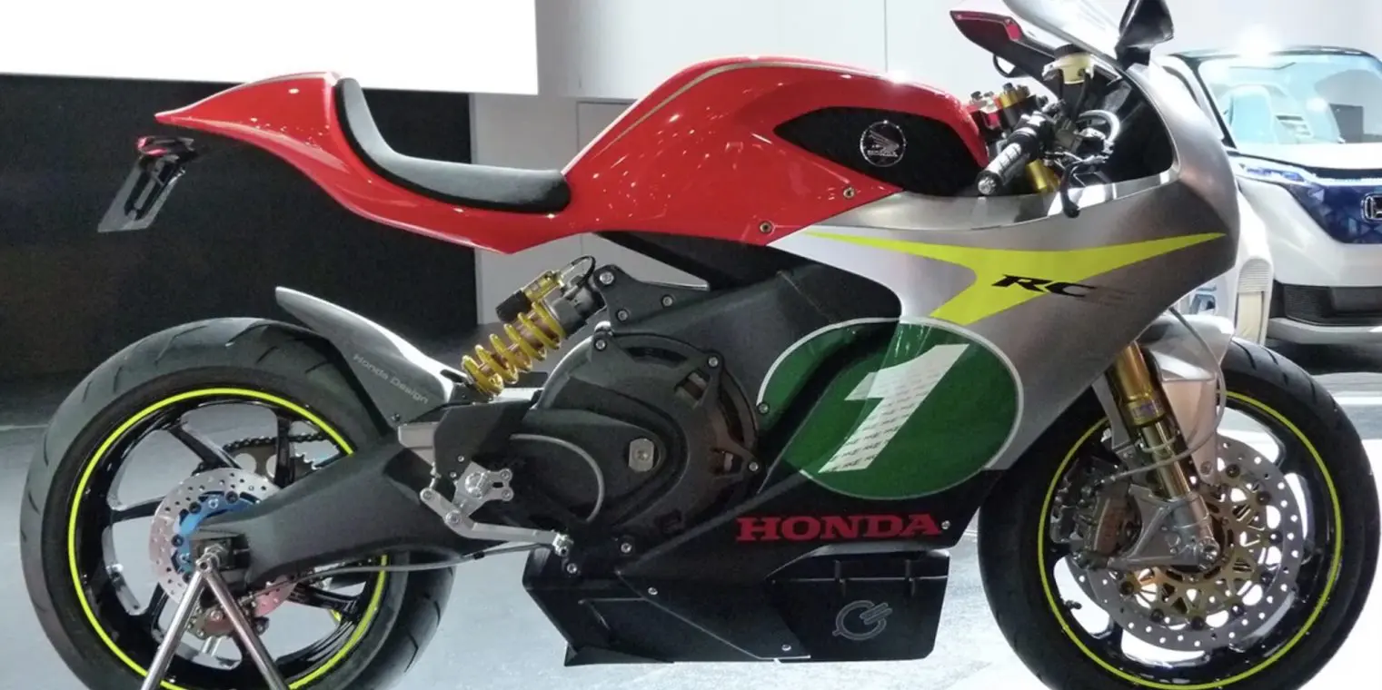 A Honda RC electric motorcycle with a red and silver body, green number plate, yellow shock absorbers, and black wheels with yellow rims, displayed indoors.