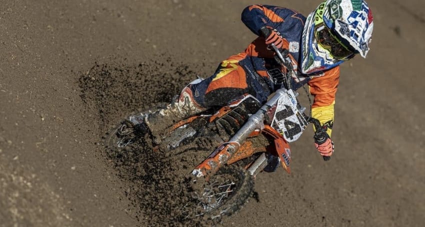 Motocross rider taking a sharp turn on a dirt track, kicking up mud.