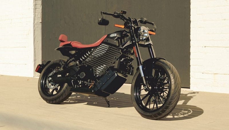 A black electric motorcycle with red seat is parked on a light-colored wooden floor against a white and dark gray wall.