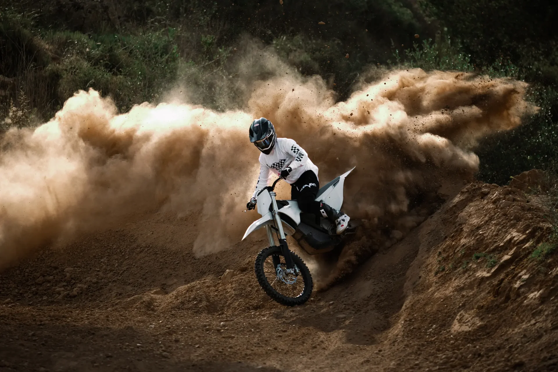 A person in a white shirt and helmet rides a dirt bike through a turn, creating a large dust cloud in the background.