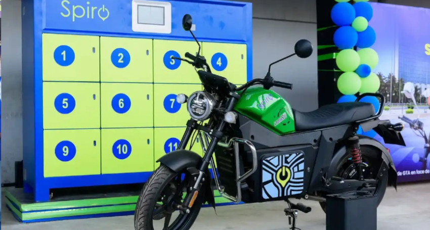 A green and black electric bike is stationed in front of a battery swapping station with numbered compartments at a Spiro facility, highlighting the convenience of seamless battery replacement.