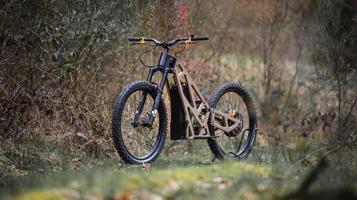 A tan-colored, off-road electric bike with thick tires and a sturdy frame is positioned on a grassy trail surrounded by dense foliage.