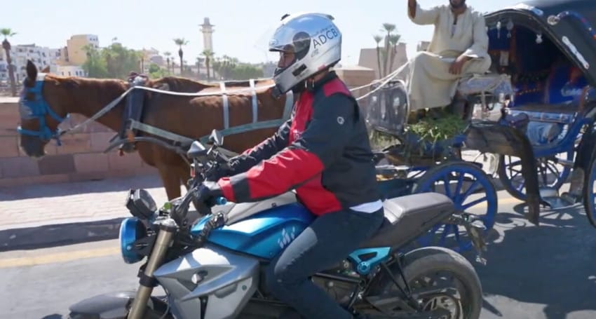 A person wearing a helmet and red jacket rides a motorcycle on a street, passing by a horse-drawn carriage.