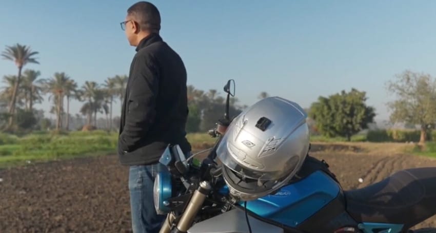 A person in a black jacket stands next to a blue electric motorcycle with a silver helmet on the handlebars, gazing at a field with trees in the background, perhaps dreaming of adventures in Egypt.