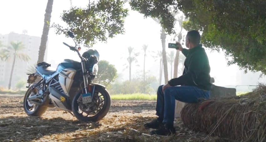 A person sits on a hay bale, capturing a photo of an electric motorcycle parked under a tree, with palm trees and buildings in the background, showcasing the blend of modern technology and timeless landscapes in Egypt.