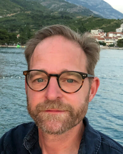 A man with glasses and a beard stands in front of a body of water with hills and buildings in the background.