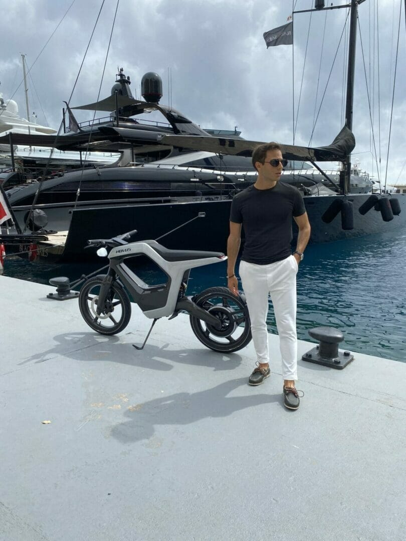 A man stands on a dock next to a sleek, modern electric bike with a large yacht in the background on a cloudy day.