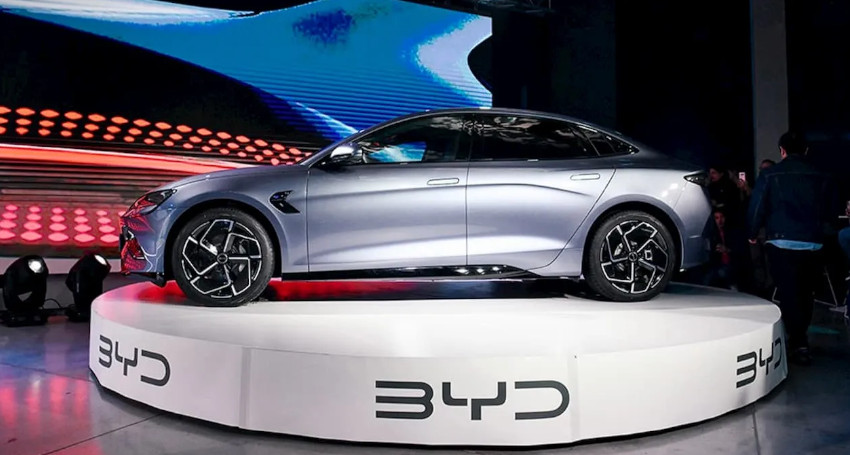 A sleek silver car is displayed on a round platform with the logo "BYD" on the base, against a backdrop of swirling light patterns.