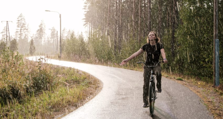 A person rides a bicycle on a winding path through a forest while it rains, with their arms outstretched and a content expression on their face.