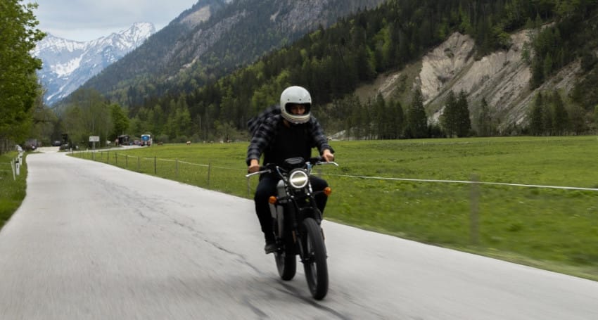 A person wearing a helmet rides a motorcycle on a road surrounded by green fields and mountains under a cloudy sky.