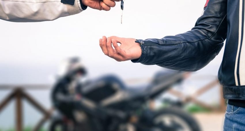 A person in a white jacket hands motorcycle keys to another person in a black jacket, with a motorcycle blurred in the background.