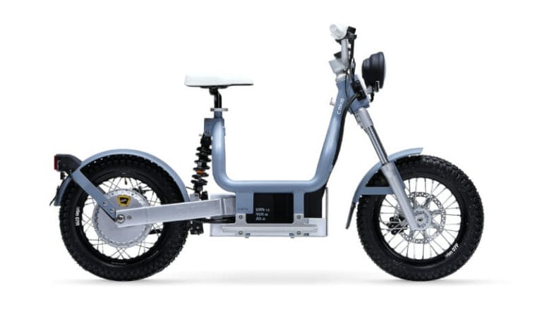 Side view of a compact electric motorcycle with a silver frame, white seat, and large knobby tires. The motorcycle has a minimalist design with visible battery compartment and rear suspension.