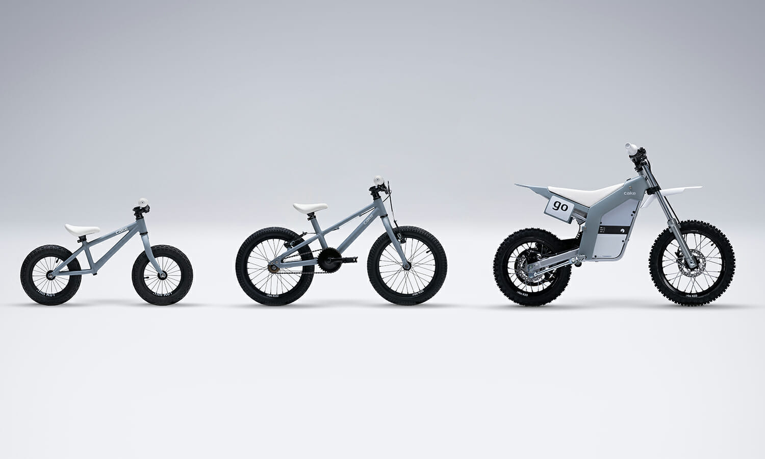 Three grey bikes are lined up from left to right, showing progression in size and complexity: a balance bike, a small pedal bike, and an electric off-road motorcycle.