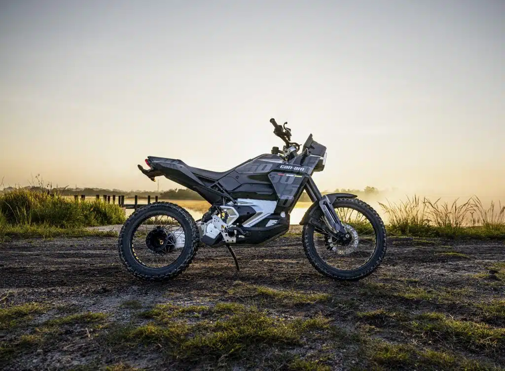 A motorcycle parked on a dirt path near a body of water during sunrise or sunset, with mist visible in the background.