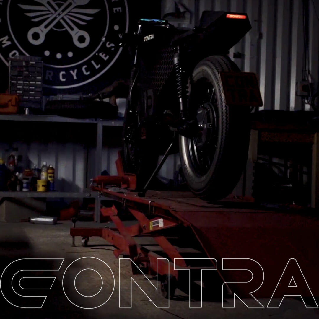 A motorcycle is positioned on a lift in a garage with the word "CONTRA" overlayed at the bottom. Tools and equipment are visible in the background.