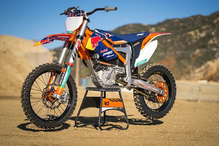 A KTM dirt bike with Red Bull branding is displayed on a stand in an outdoor, mountainous setting.