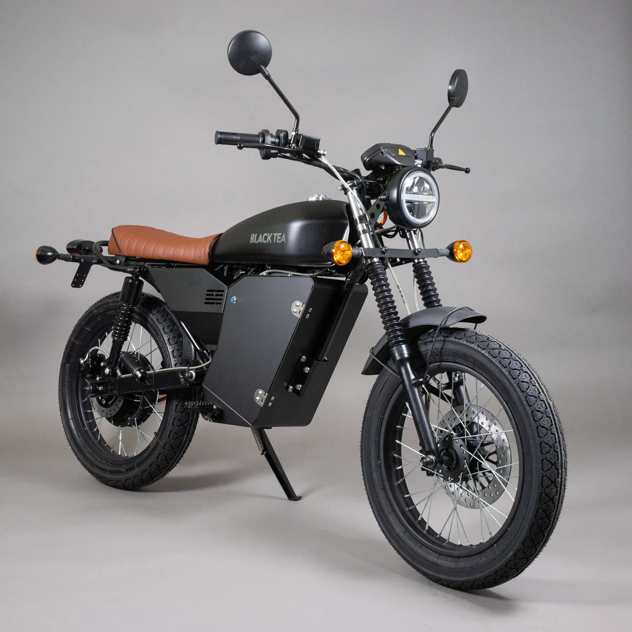 A black electric motorcycle with a brown seat, round headlight, and side mirrors against a plain gray background. The text "Black Tea" is visible on the side of the tank.