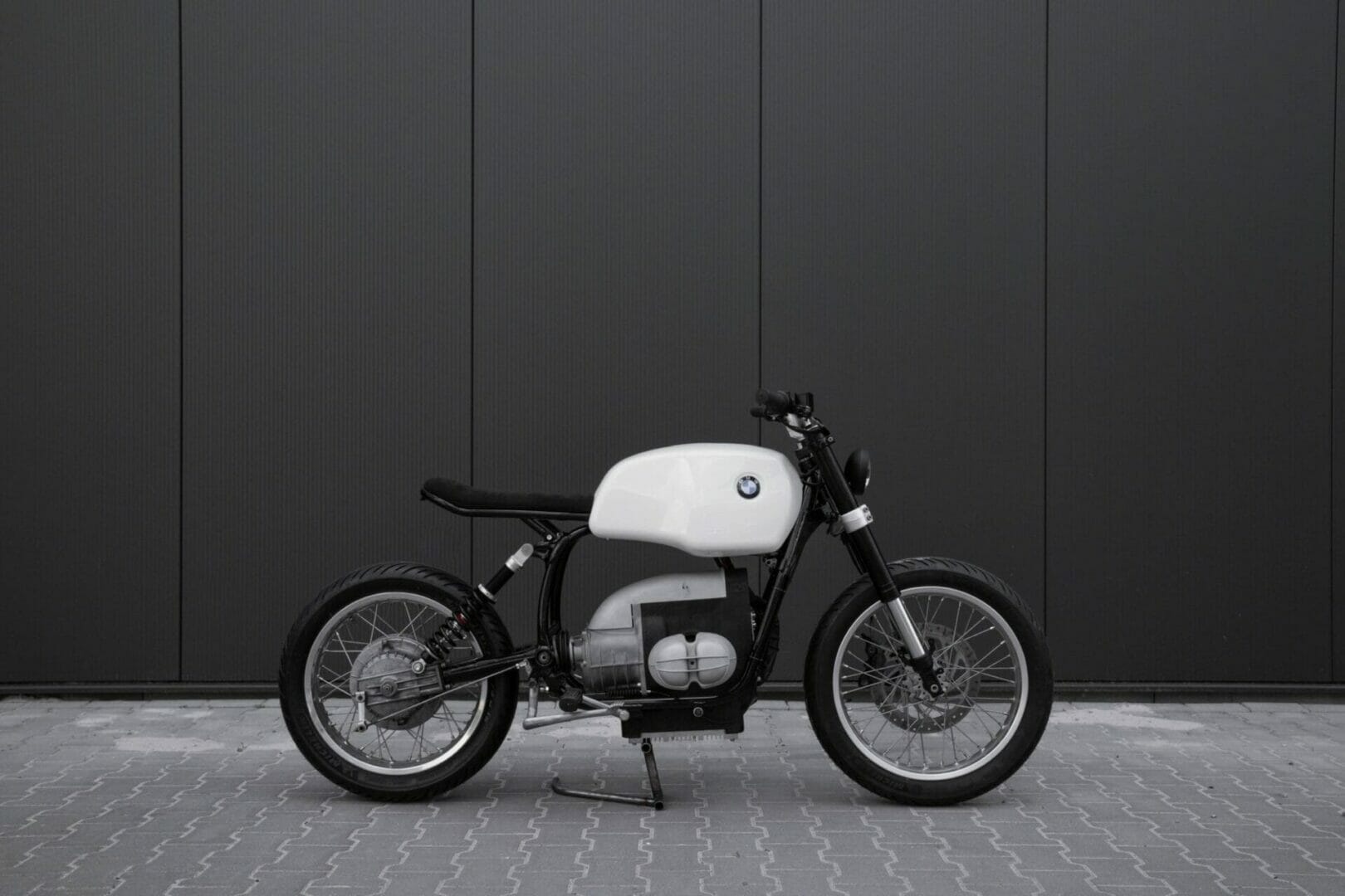 A minimalist custom-built motorcycle with a white fuel tank and black frame is parked on a tiled surface against a dark background.