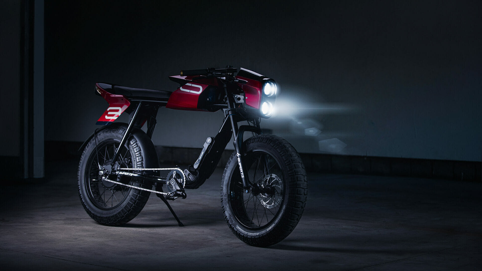 A red and black electric motorcycle with large tires and dual headlights is illuminated in a dark setting.