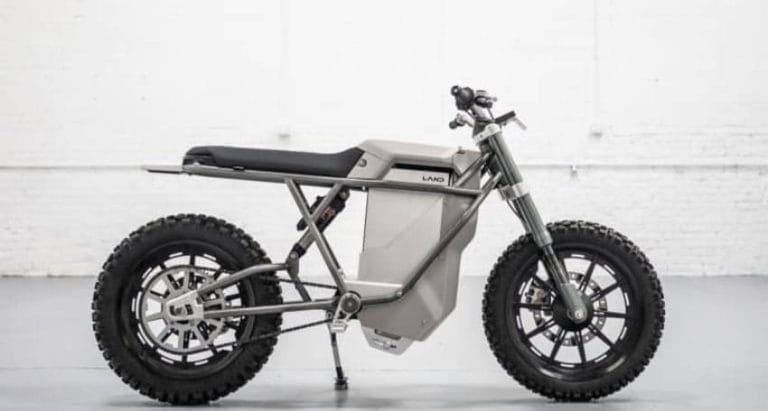 A minimalist grey electric motorcycle with a rugged build, off-road tires, and a geometric frame is displayed against a white industrial background.