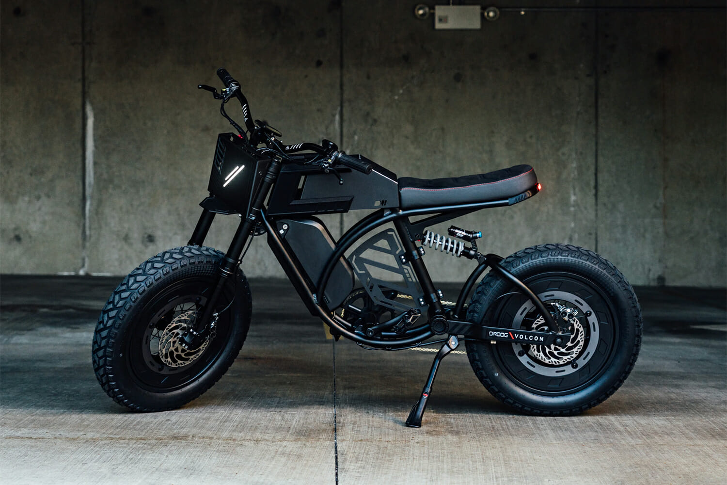 A black electric bike with thick tires, a minimalist frame, and a padded seat is parked indoors against a concrete wall.