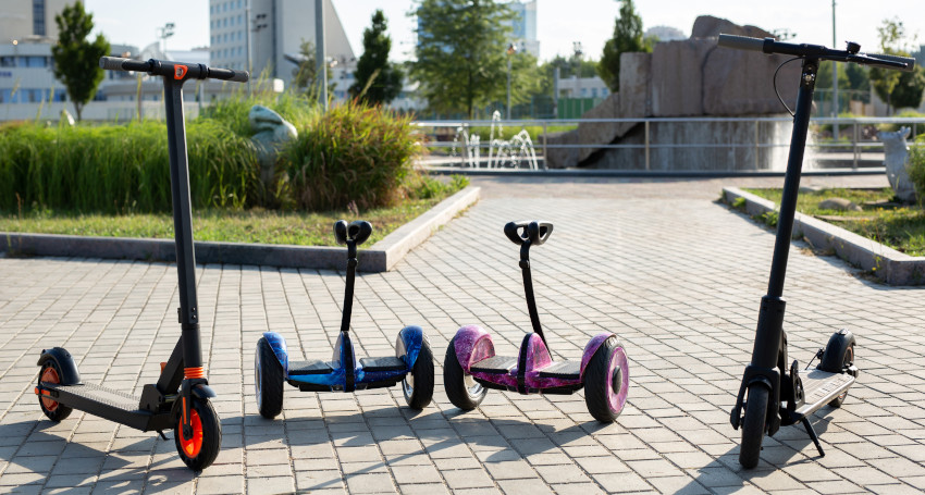 Four electric scooters and balance boards are parked on a paved path in an outdoor urban setting.