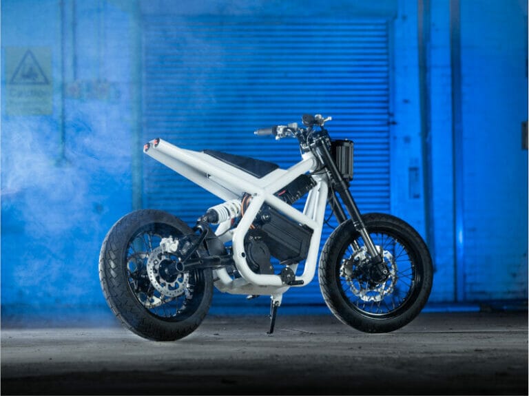 A white electric motorcycle is parked in an industrial setting with a blue-lit background and slight fog, showcasing its design and structure.