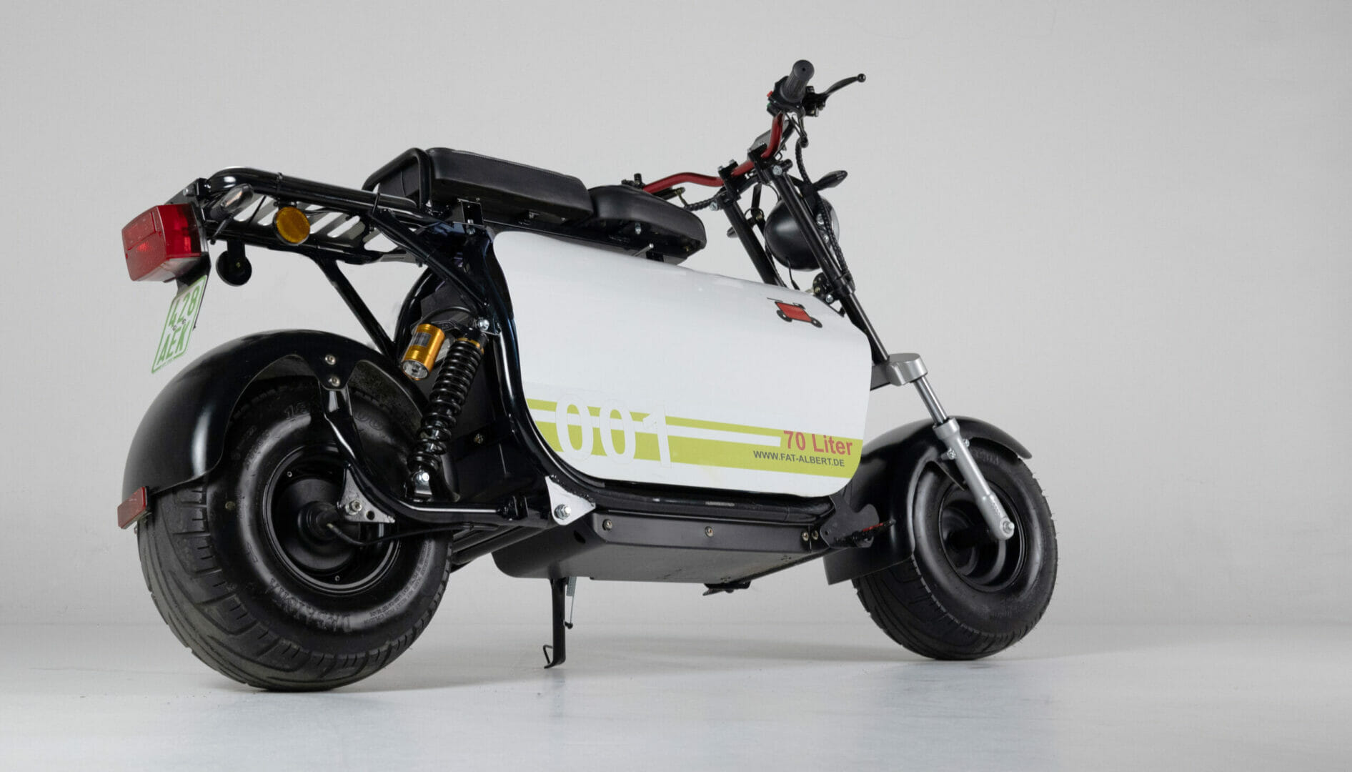 A side view of an electric cargo scooter with a white frame, black seat, black handlebar, and large tires. The rear wheel has a red reflector and there is a green license plate on the left.