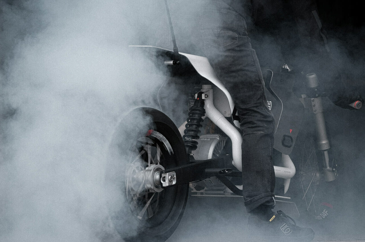 A person wearing dark jeans performs a burnout on a motorcycle, causing thick white smoke to billow from the rear tire.