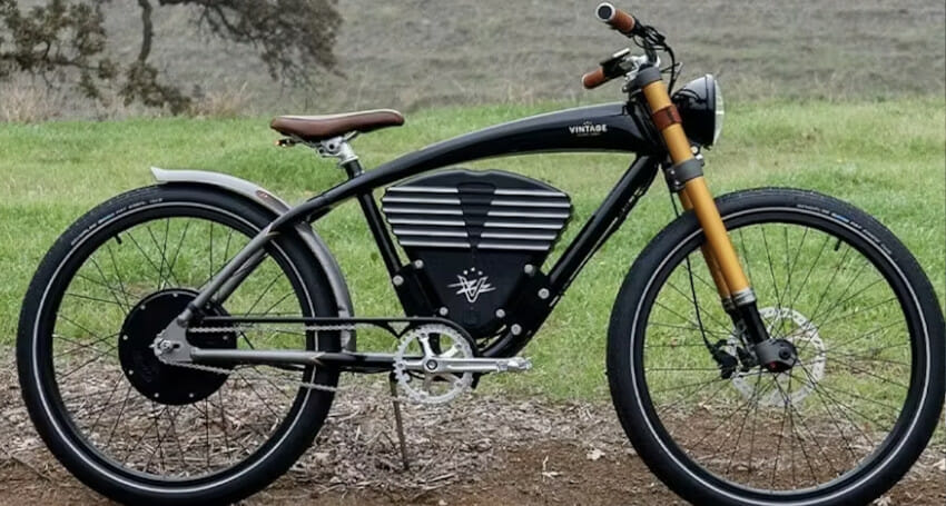 A black electric bike with vintage design elements is parked on a grassy area with a forest background.