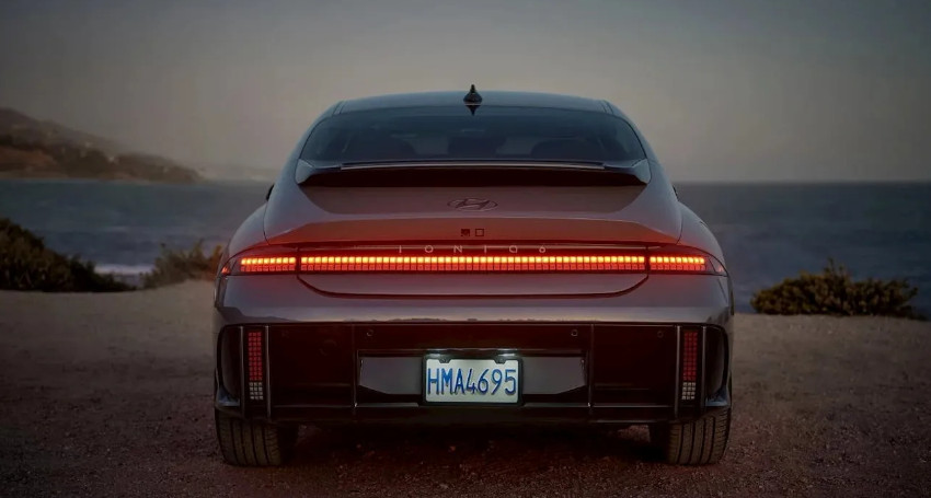 A dark-colored Hyundai IONIQ 6 electric car is parked by the coastline at dusk, with its rear facing the camera. The license plate reads "HMA 4695" and the taillights are illuminated in a horizontal strip.