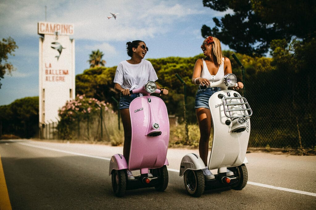 Two women riding scooters down a road.