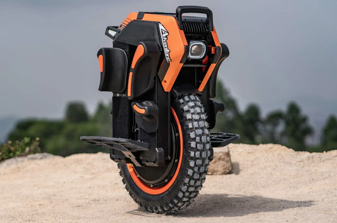 Orange and black electric unicycle with large treaded tire, resting on sandy terrain against a blurred outdoor background.