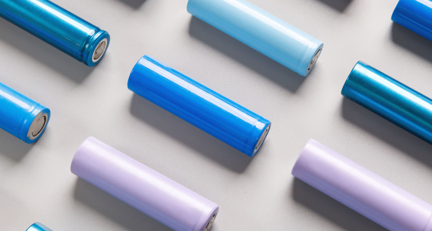 Rows of cylindrical lithium-ion batteries in various shades of blue, purple, and light blue arranged on a light grey surface.