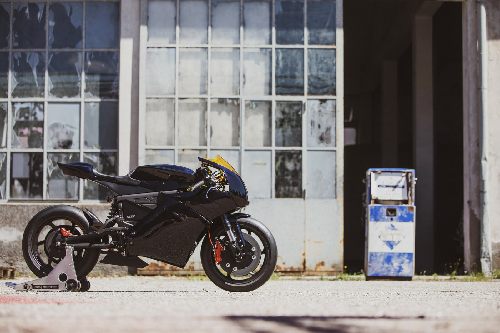 A black sports motorcycle is parked in front of an old industrial building with large windows and beside a weathered blue and white fuel pump.