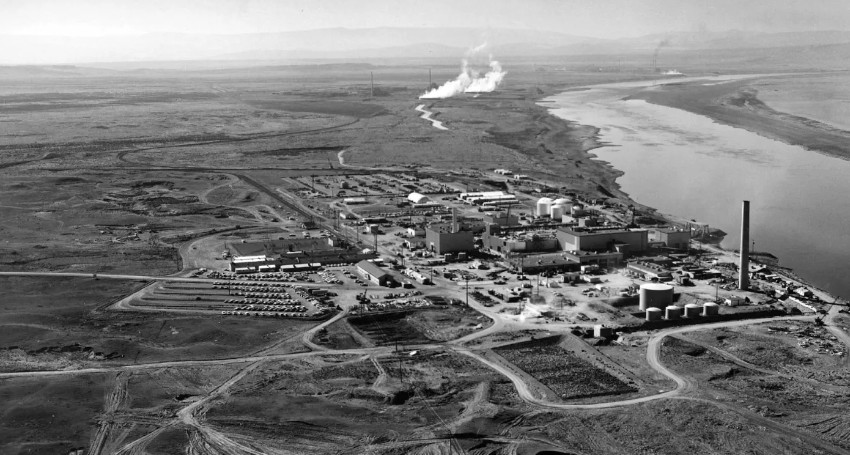 Black and white aerial view of an industrial site near a river, with buildings, storage tanks, parking lots, and billowing smoke in the background. Sparse landscape surrounds the facility.