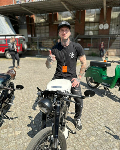 A man sits on a parked motorcycle, wearing a black t-shirt, black pants, and a cap. He is giving a thumbs-up. Other motorcycles and people are visible in the background.