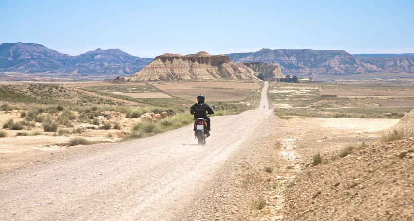 A person rides a motorcycle on a deserted, gravel road through an arid landscape with distant mountains and rock formations under a clear blue sky.