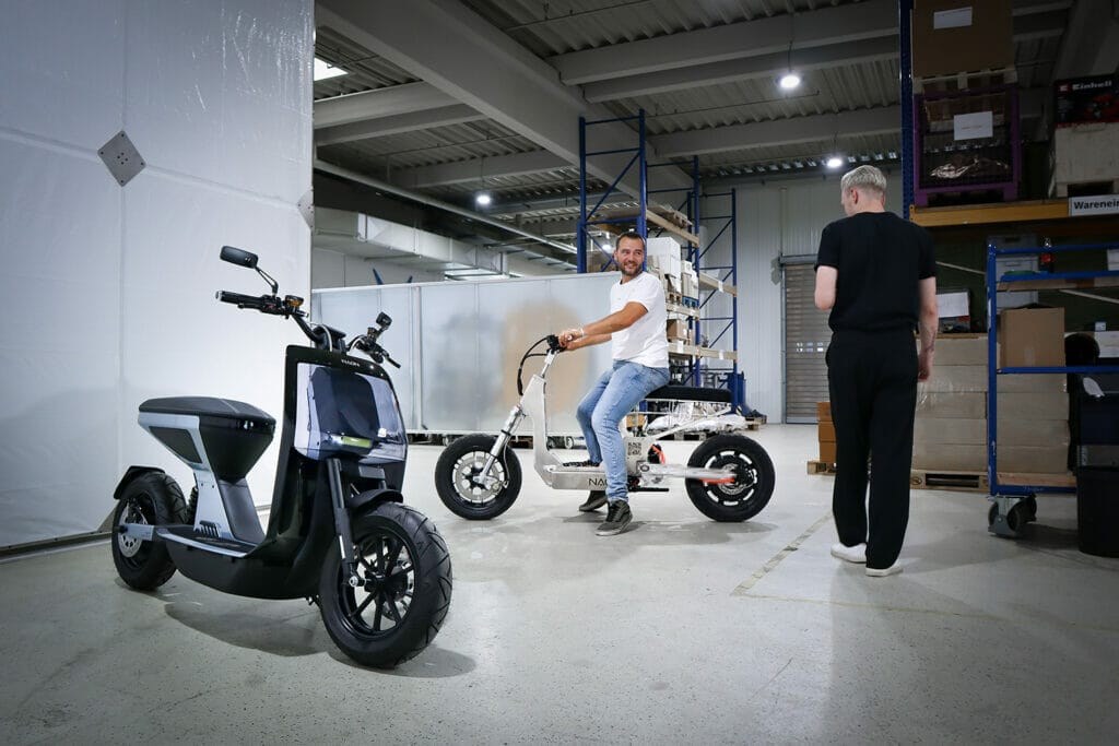 Two men standing next to two electric scooters in a warehouse during an Auto Draft event.