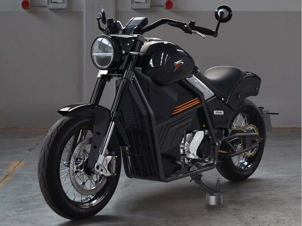 A black electric motorcycle is parked in an indoor garage. The bike has a modern design with a round headlight, angular body panels, and rear suspension.